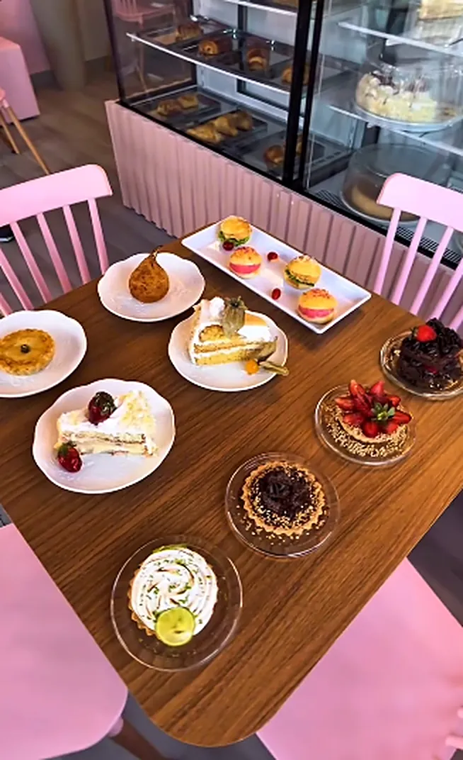 Mesa com vários doces: macarons, fatias de bolo, tortas e morangos, em frente à vitrine